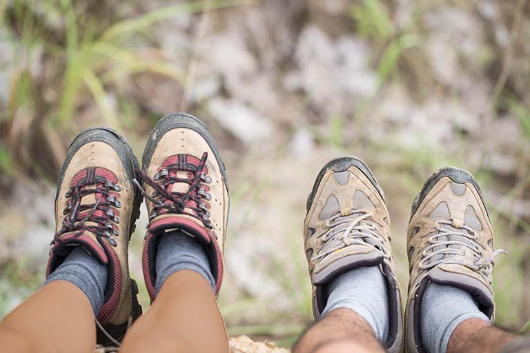 Background of blurred grass and brown leaves. In the foreground are a woman and a man's legs and feet.  Both are wearing hiking boots.  They appear to be sitting on elevated ground with their feet extending out over the edge.