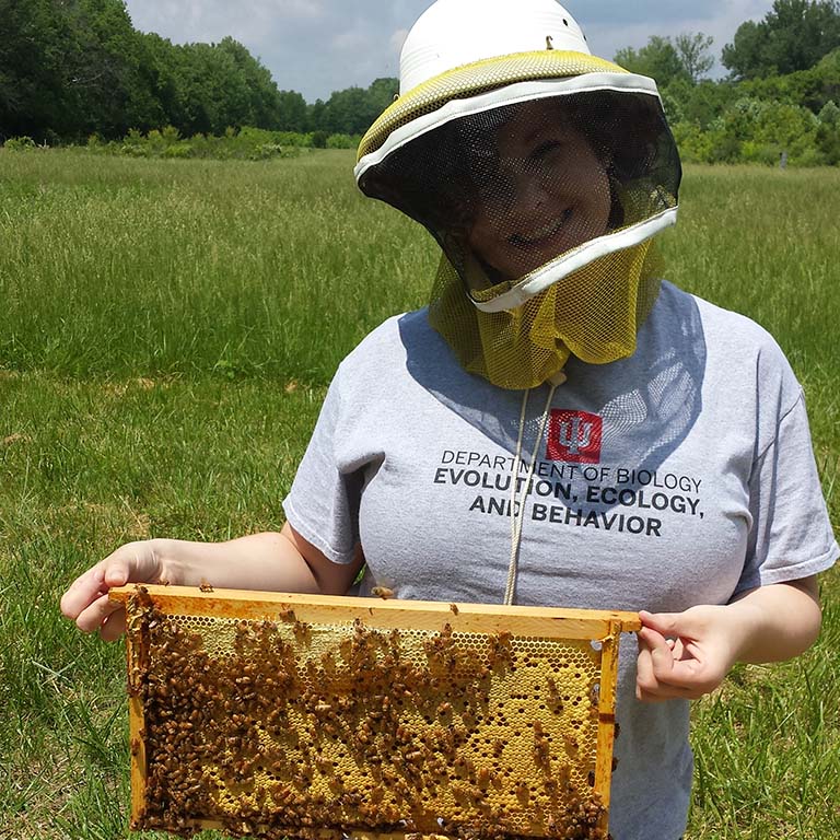Delaney Miller, wearing a beekeeper's hat with netting, presents a frame from a bee hive with honeycomb and lots of bees.