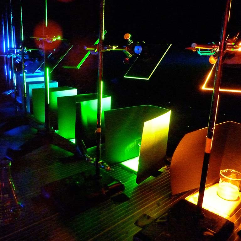 Mirrors are used to reflect different colors of light onto beakers with samples of the ocean bacterium Synechococcus under an experiment overseen by researchers from IU.
