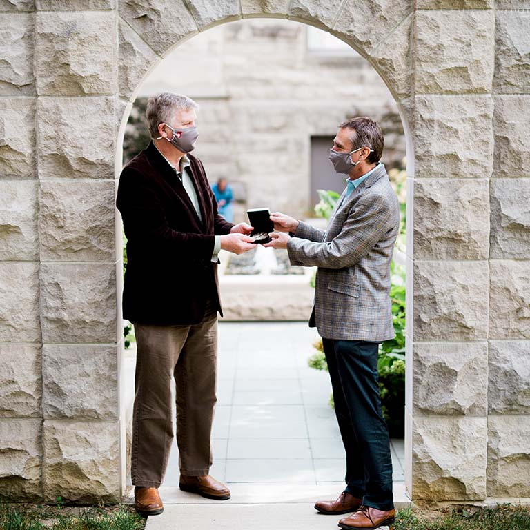 Rick Van Kooten presents Craig Pikaard (left) with the IU Bicentennial Medal. Both are standing outdoors in a stone archway on the IU Bloomington campus.