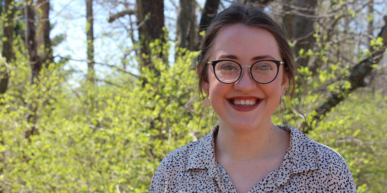 Abigail McClain poses for a photo while standing in a forest; tree leaves are beginning to appear.