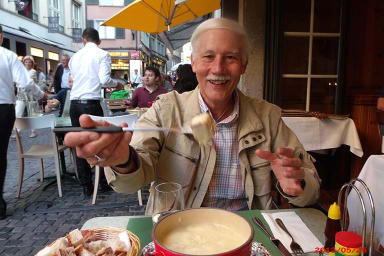 Alum Michael Miller offers a bite of food toward the camera photographing him at an outdoor dining table in Switzerland.