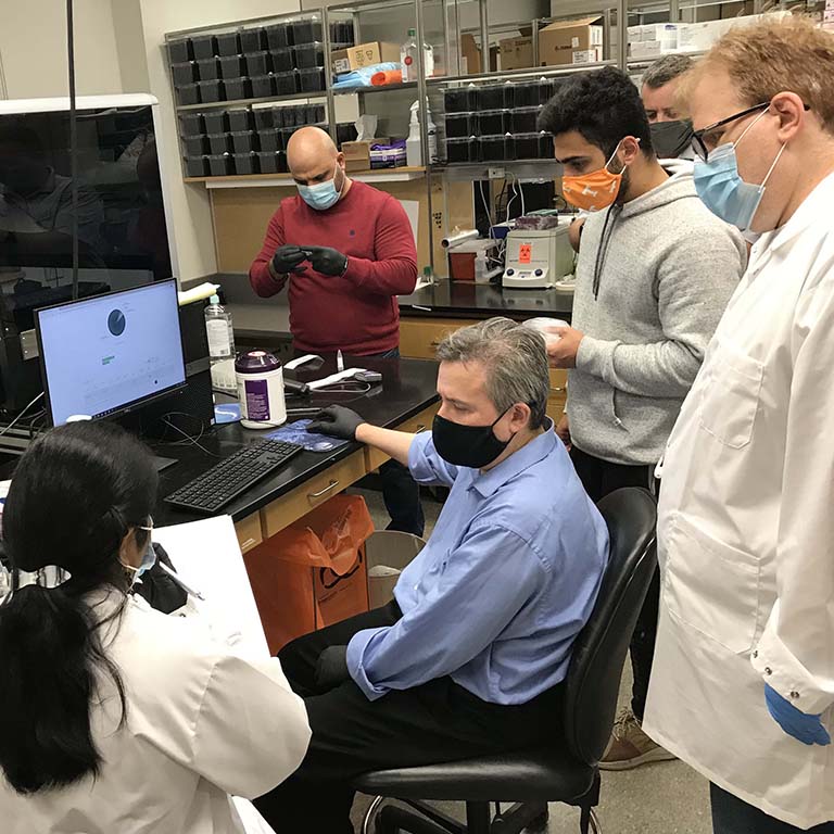 Six masked and gloved team members gather around the computer, evaluating the first test results at 8:15 p.m. on the evening of the facility's first day, October 21, 2020.