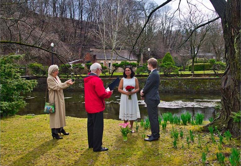 Amrita Bhattacharya and Jon Zarling stand in a lovely outdoor setting with pond and greening shrubs and grass as the daffodil foliage pushes through the ground on March 20, 2020, in Pennsylvania during their civil wedding ceremony.